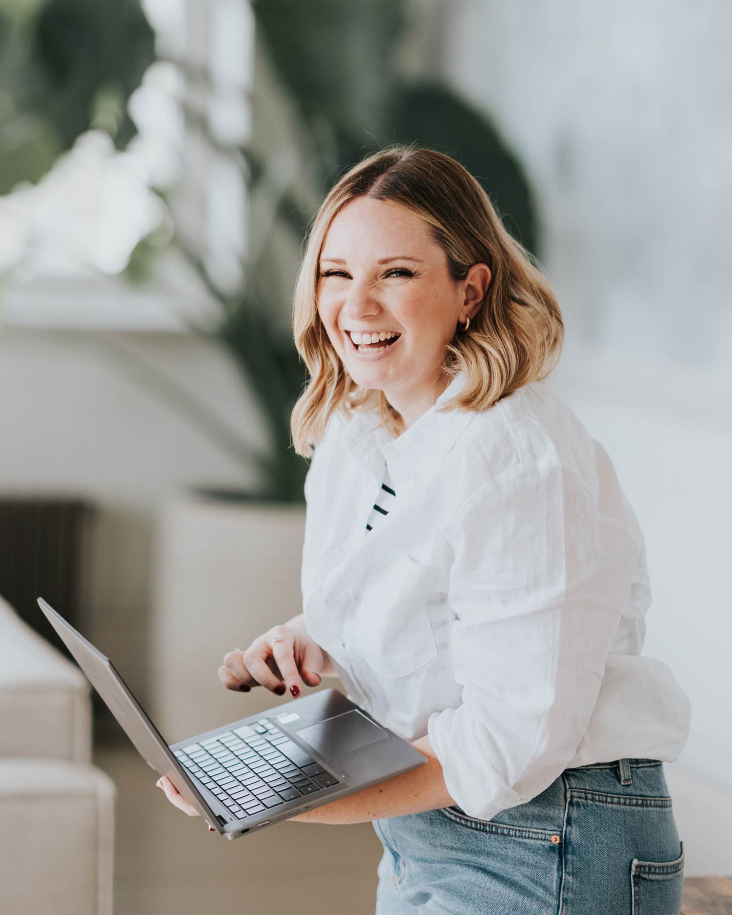 Person in white shirt laughing while using laptop in bright room with plants.