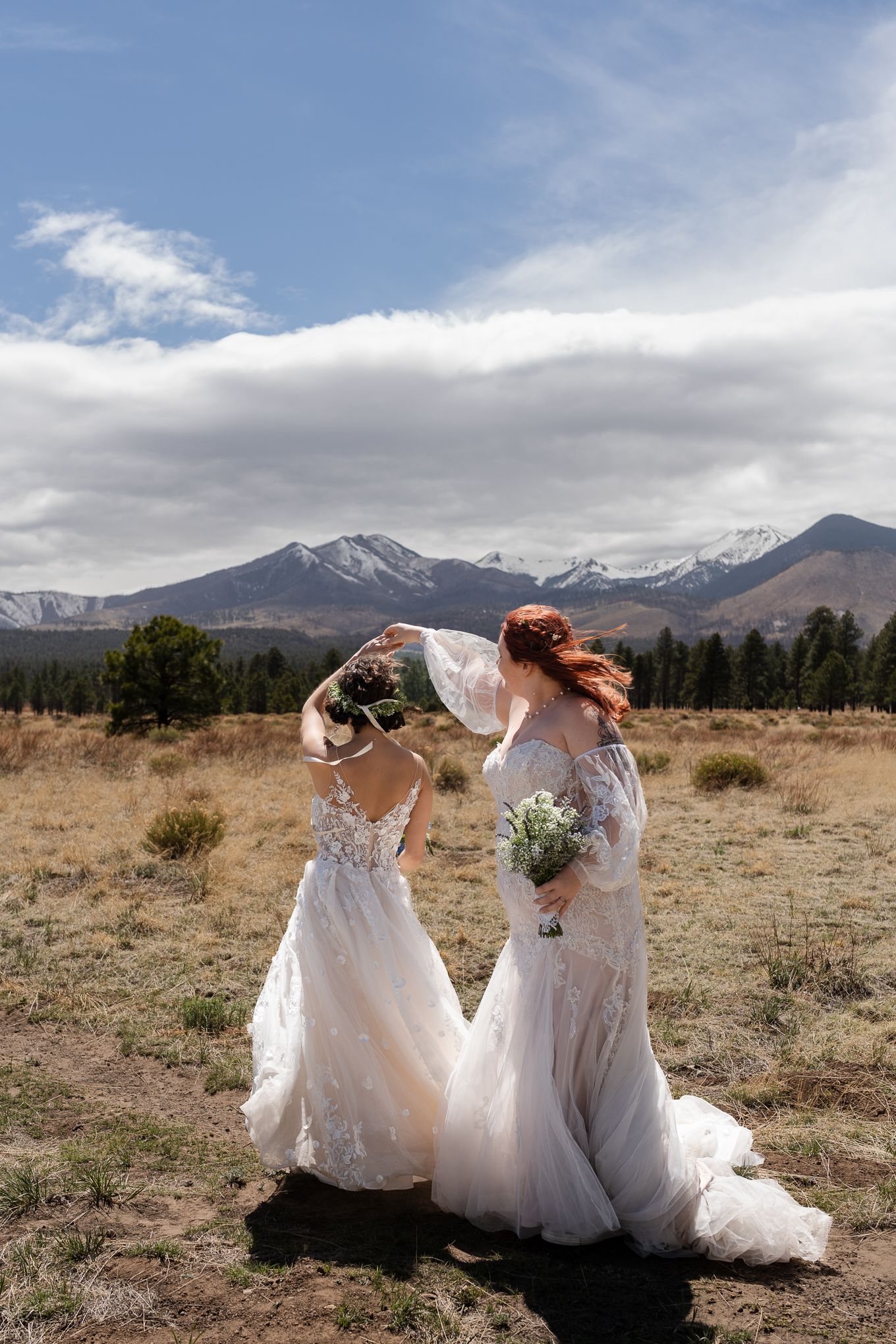 Two brides in white lace dresses twirl together against a mountain landscape.