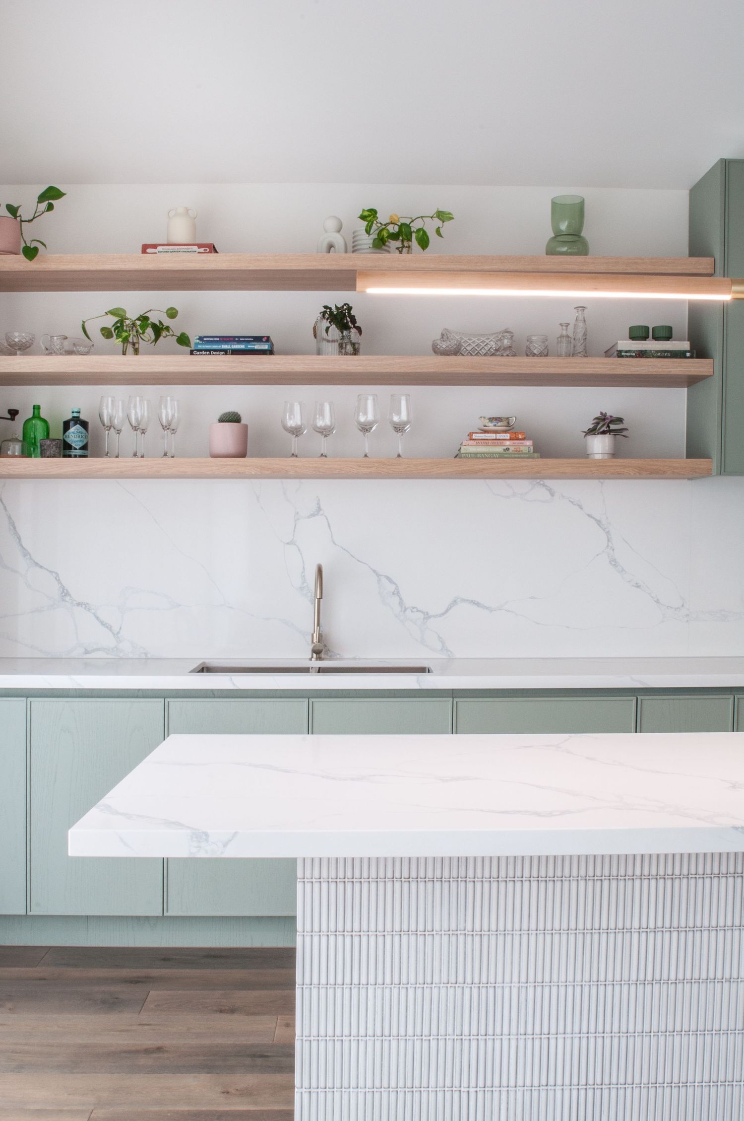 Kitchen with sage green cabinets, marble backsplash, and wooden floating shelves displaying glassware.