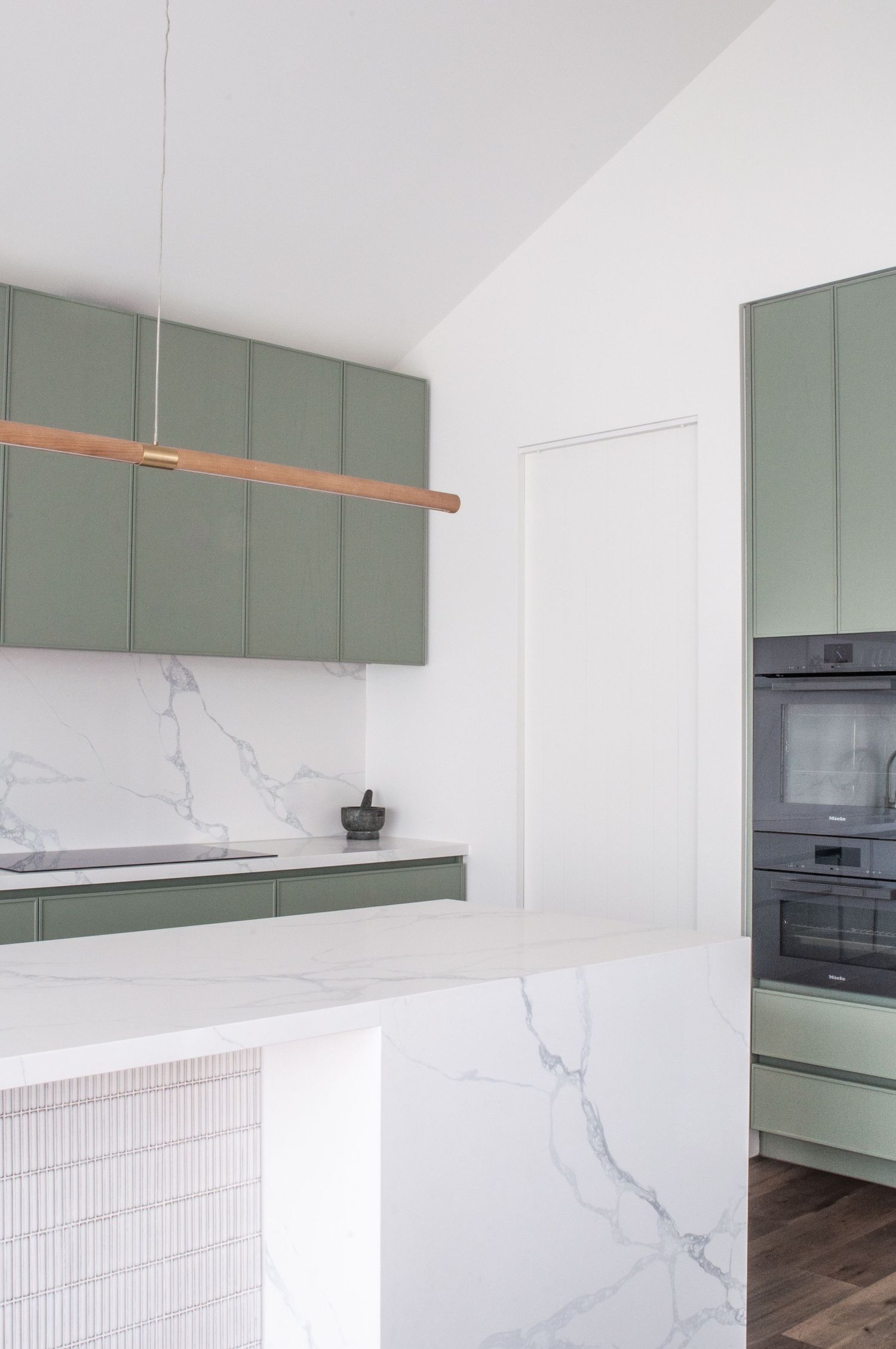 Modern kitchen with sage green cabinets, marble countertops and suspended wooden light fixture.