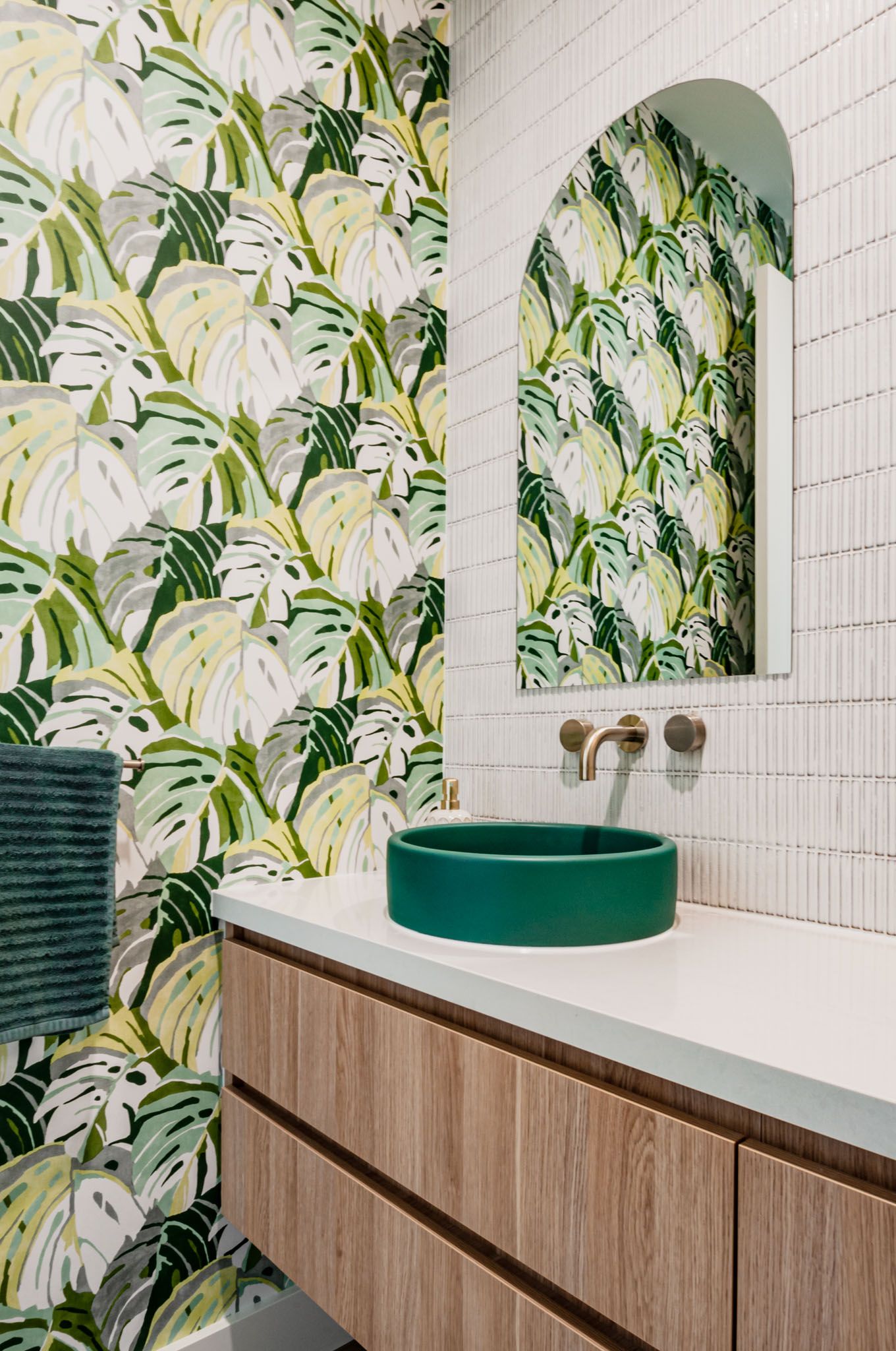 Trendy bathroom with tropical leaf wallpaper, green vessel sink, and wooden vanity against white tile walls.