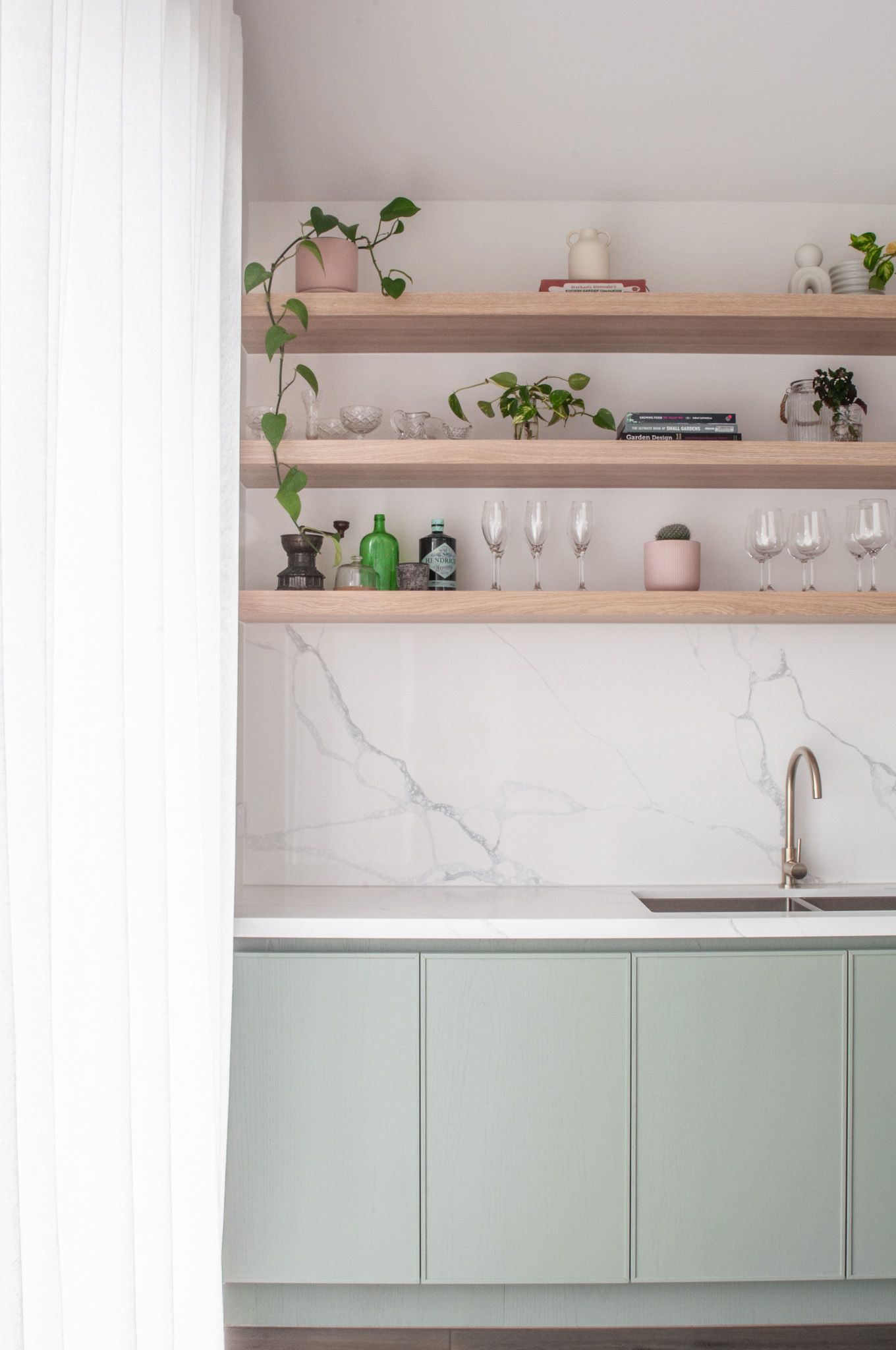 Modern kitchen nook with wooden shelving, marble backsplash, and sage green cabinets displaying glassware.