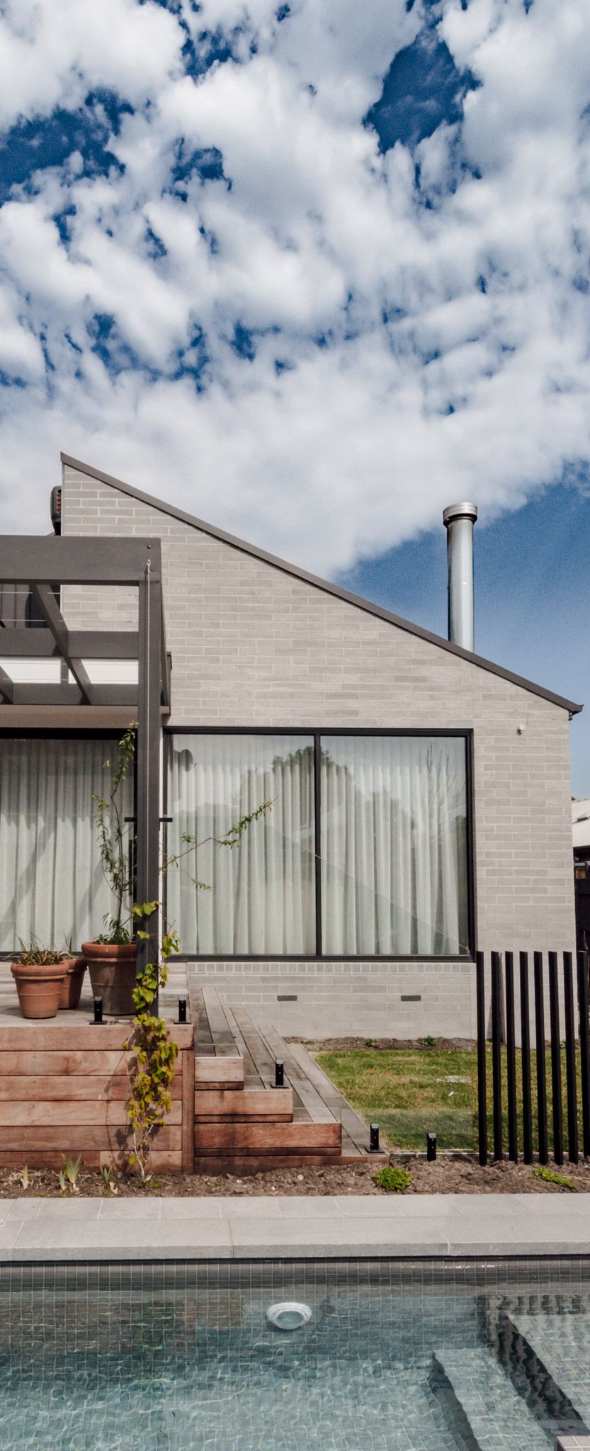 Modern poolside view of a minimalist house with brick exterior, wooden steps, and potted plants.