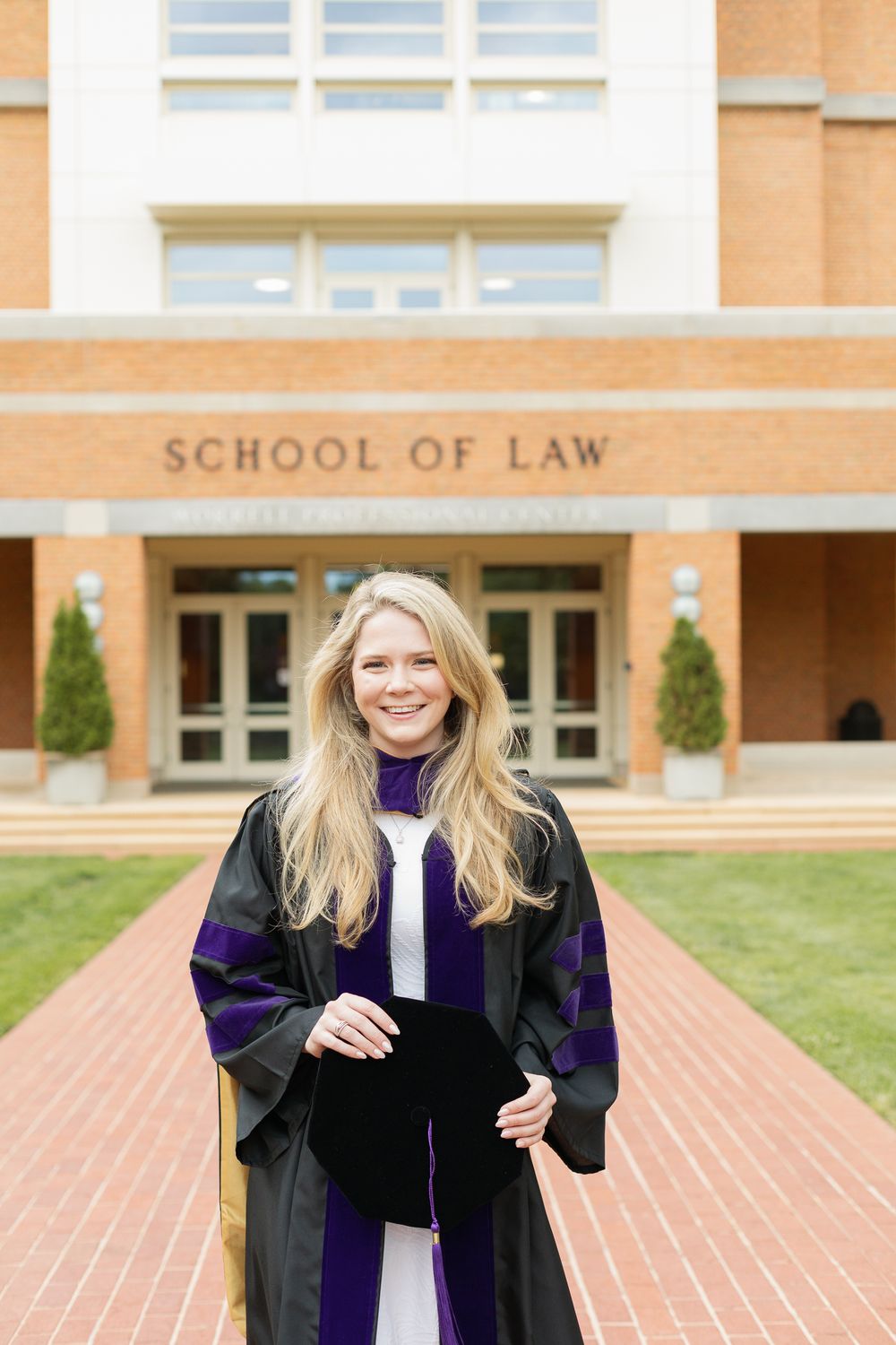 A graduate in academic regalia poses proudly outside the School of Law building.