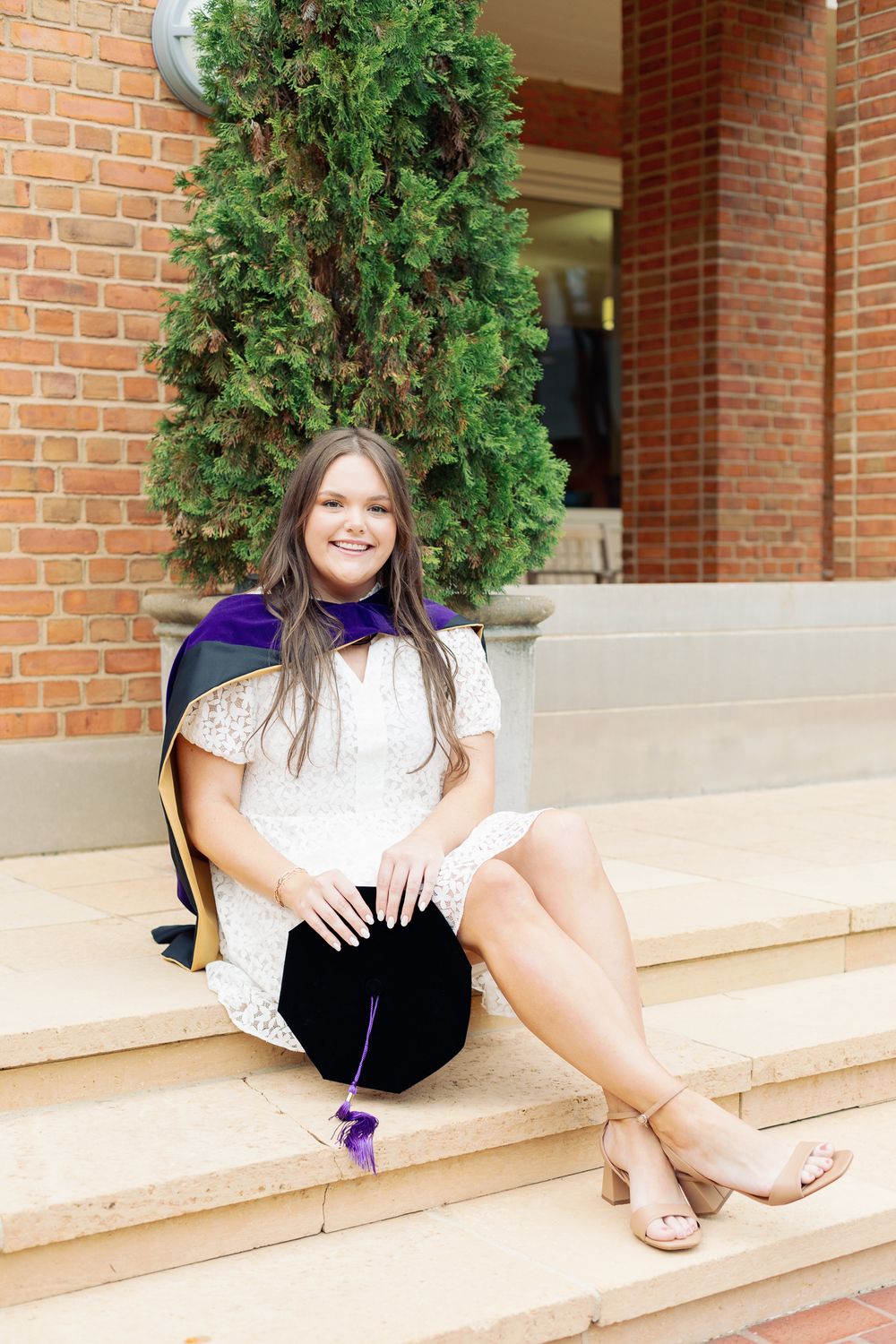 Graduate in white dress and graduation stole relaxes on concrete steps by brick columns.