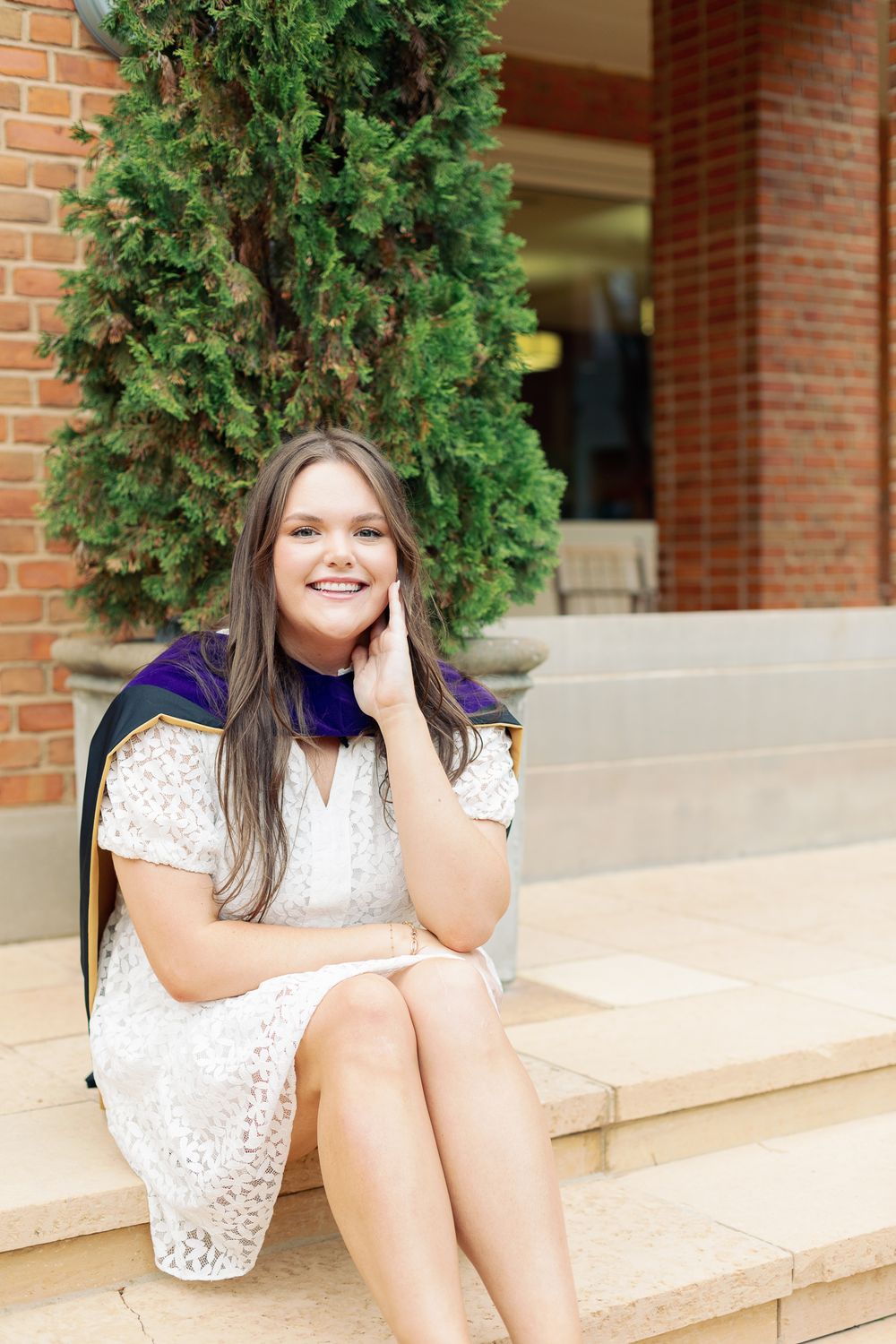 Graduate in lace dress poses casually on outdoor campus steps.