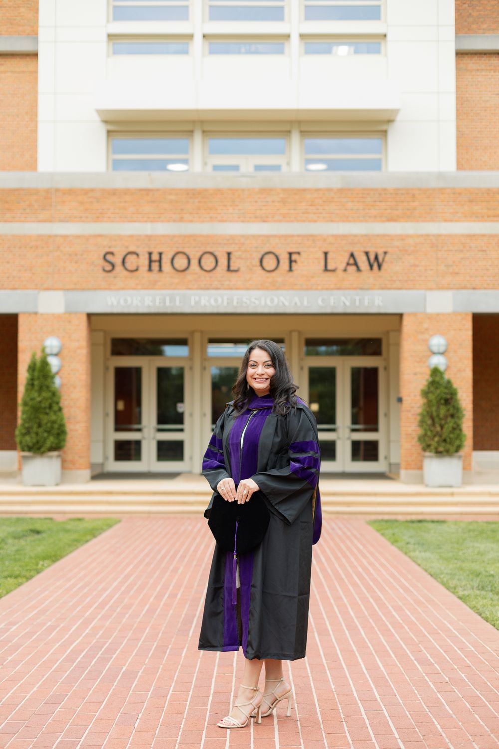 Graduate in academic regalia poses confidently outside law school entrance.
