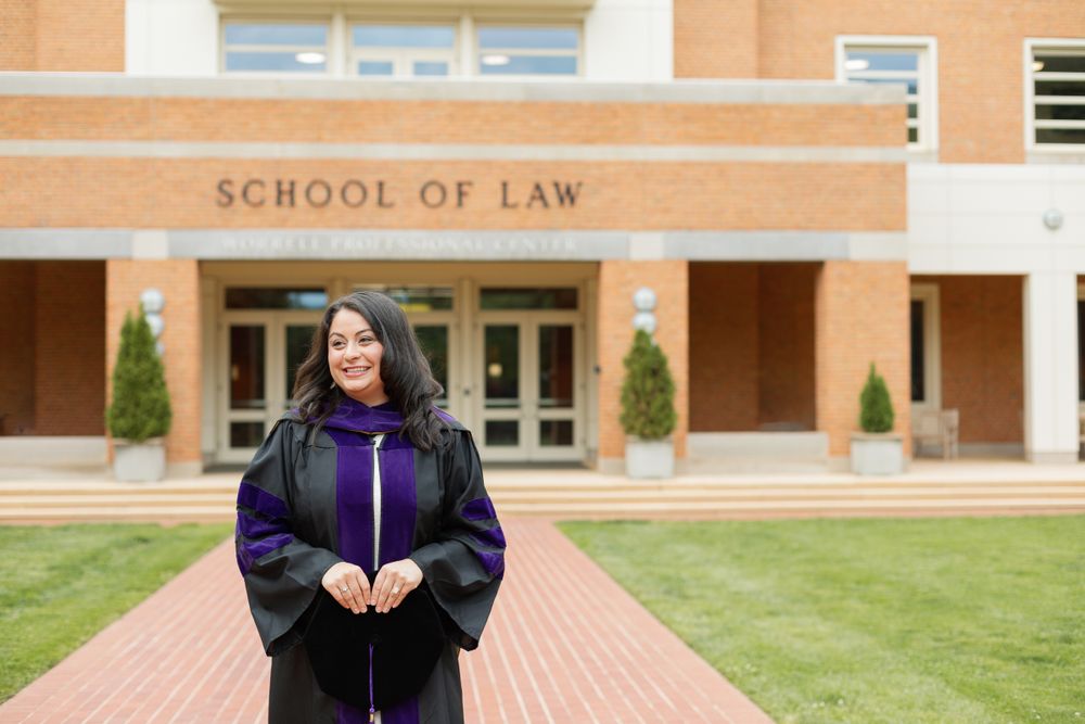 Graduate in black and purple regalia stands in front of School of Law sign.