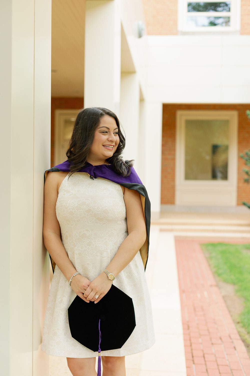 Graduate in white sleeveless dress poses elegantly in columned hallway.