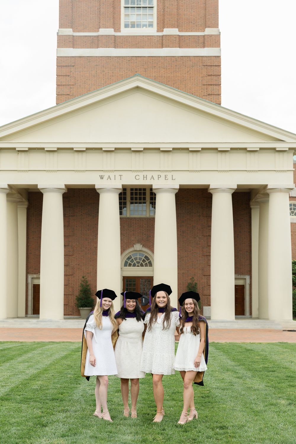 Graduates in matching white dresses gather on lawn in front of chapel's columned entrance.