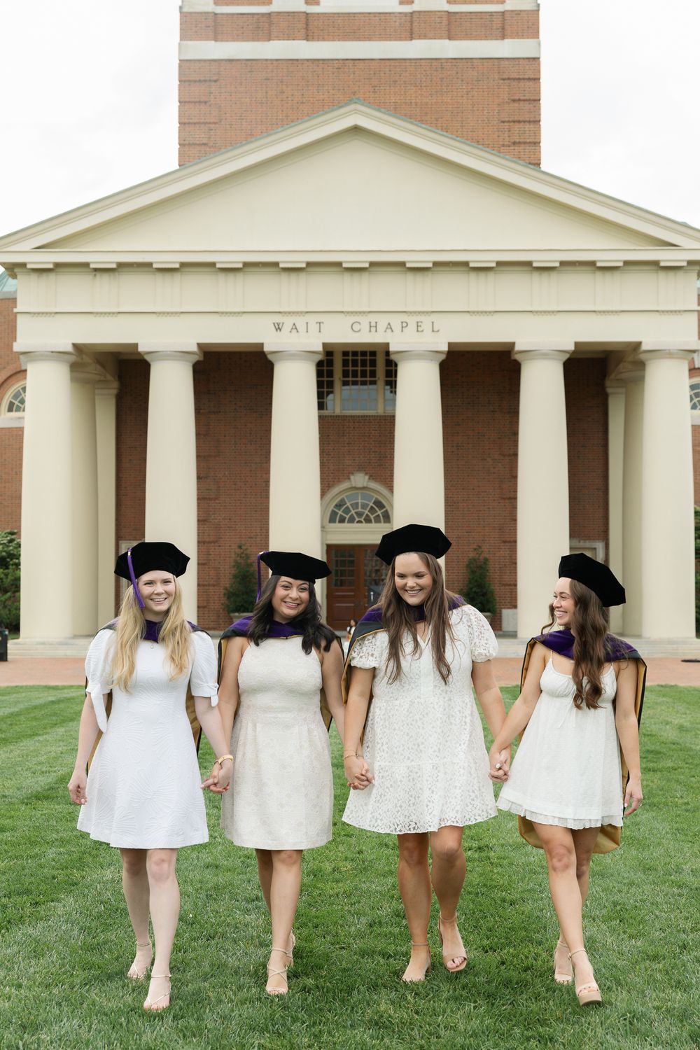 Graduates in white dresses walk together across lawn in front of chapel architecture.