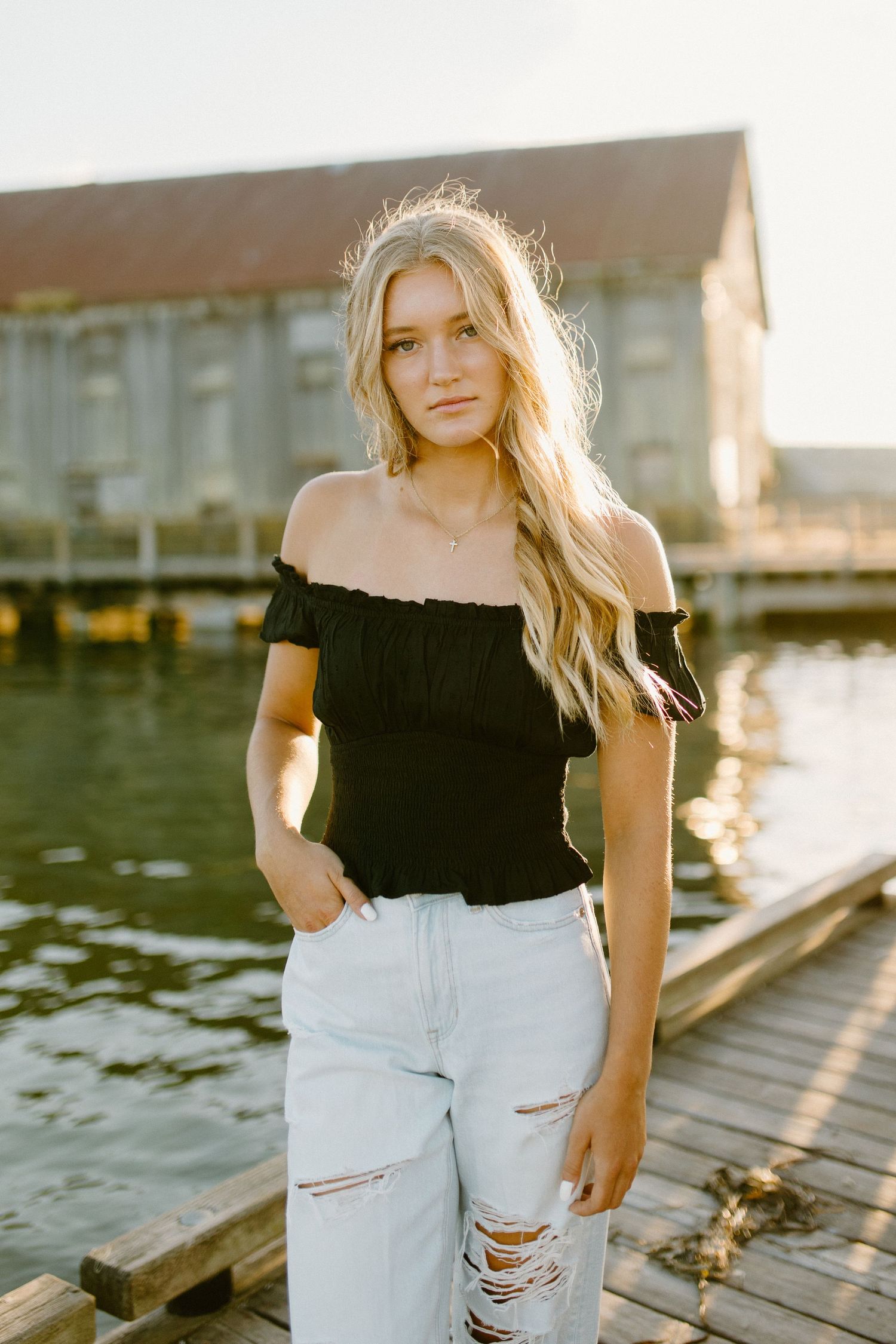 Woman in black off-shoulder top and ripped jeans poses on wooden dock at sunset.