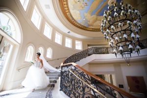 A bride posing on an elegant curved staircase beneath a grand chandelier and domed ceiling.
