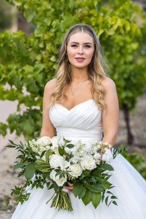 A bride in a white strapless gown holds a lush white and green bouquet against a vineyard backdrop.