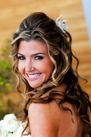 Bride with curled blonde hair and white flower accessory smiling outdoors.