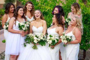 A joyful wedding party of bridesmaids in white dresses laugh together while holding white floral bouquets outdoors.