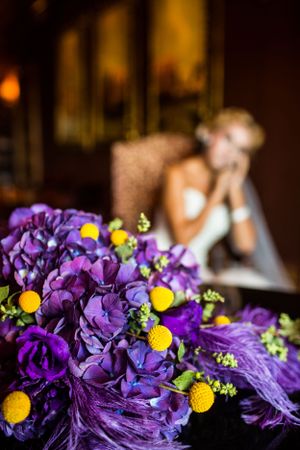 A vibrant purple and yellow wedding bouquet with feather accents in the foreground.