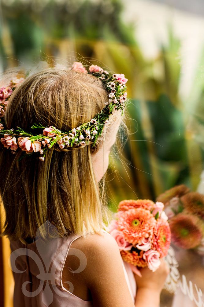 Young attendant wearing floral crown holds pink flowers in garden setting.