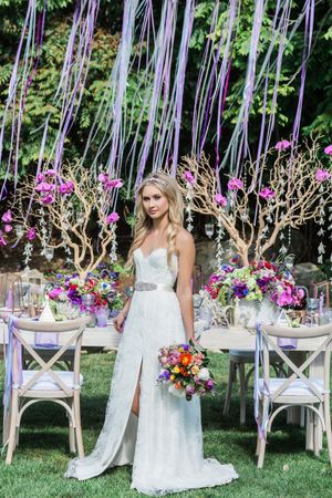 Garden wedding setup with purple ribbons and floral arrangements on rustic white chairs.