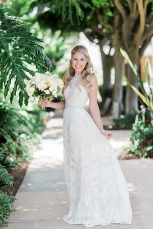 Elegant bride in lace gown holds white bouquet while posing in garden path with tropical foliage.