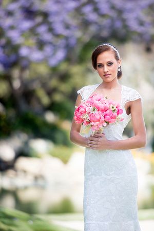 Bride in lace cap sleeve dress holds vibrant pink peony bouquet against purple floral background.