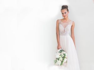 Elegant bride in flowing white dress holding white floral bouquet against bright background.