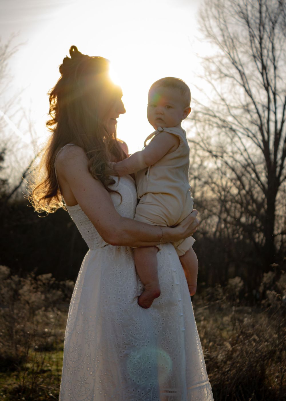 Mom in white dress holding baby with sun shining behind her