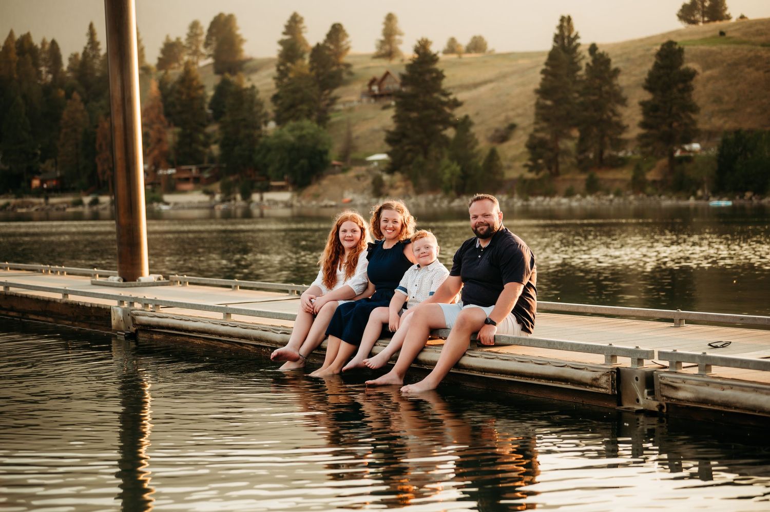 People relaxing on a wooden dock by a tranquil lake surrounded by evergreen trees and rolling hills at sunset.