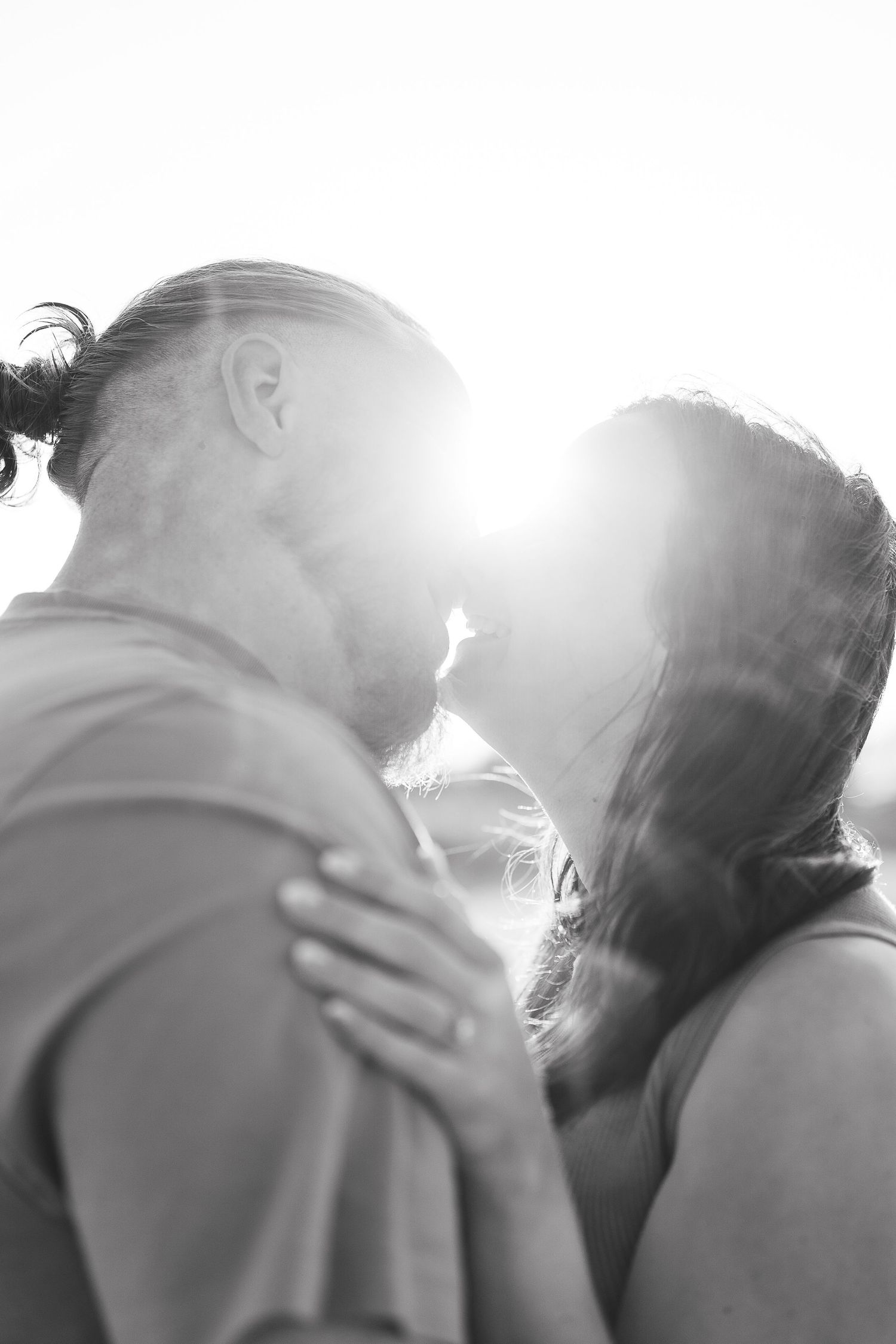 Black and white photo of a couple sharing a tender kiss backlit by bright sunlight.