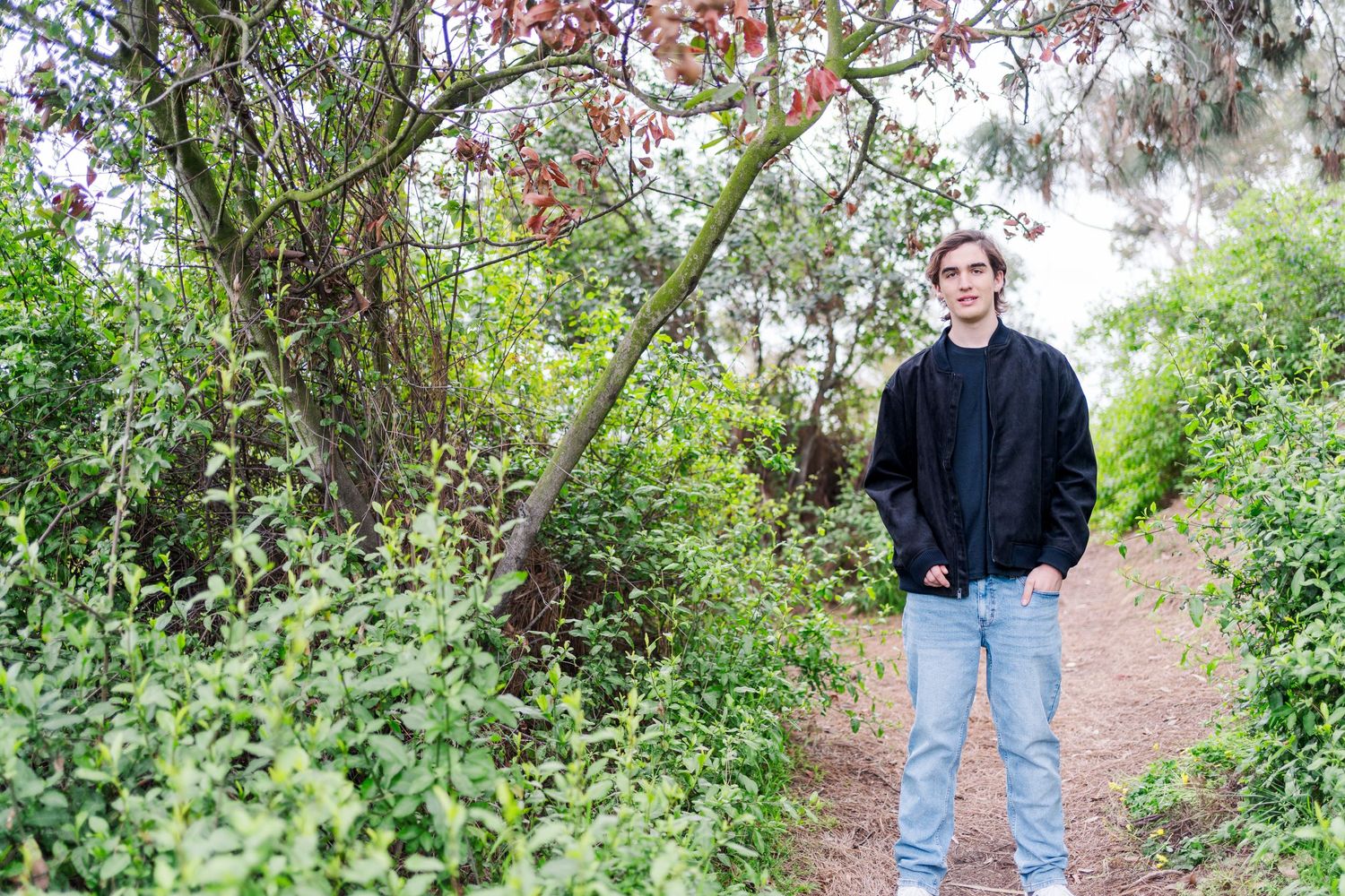 A person in a black jacket and jeans stands on a nature path surrounded by lush green foliage and pink blooming trees.