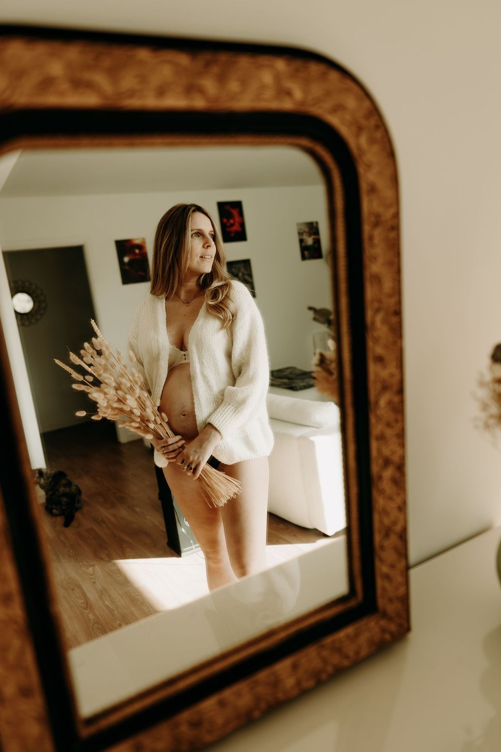 Mirror selfie reflection showing person in white sweater holding dried flowers in vintage framed mirror against dark wall.