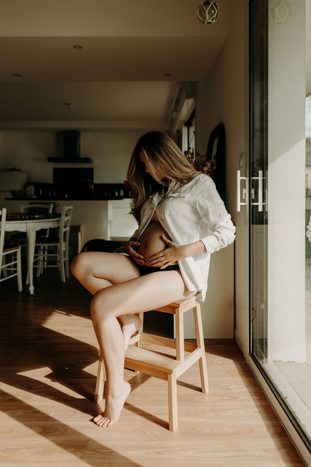 Sunlit artistic indoor portrait of a person wearing an oversized white shirt while sitting on a wooden stool.
