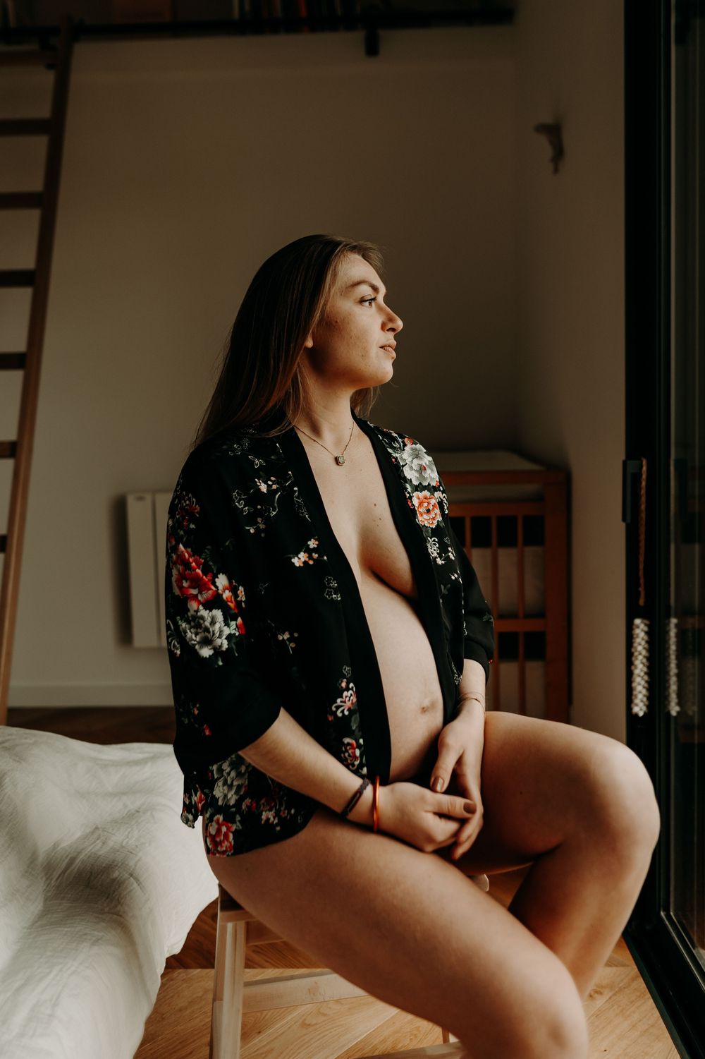 A pregnant woman sits by a window wearing a floral black robe in an intimate boudoir-style portrait.