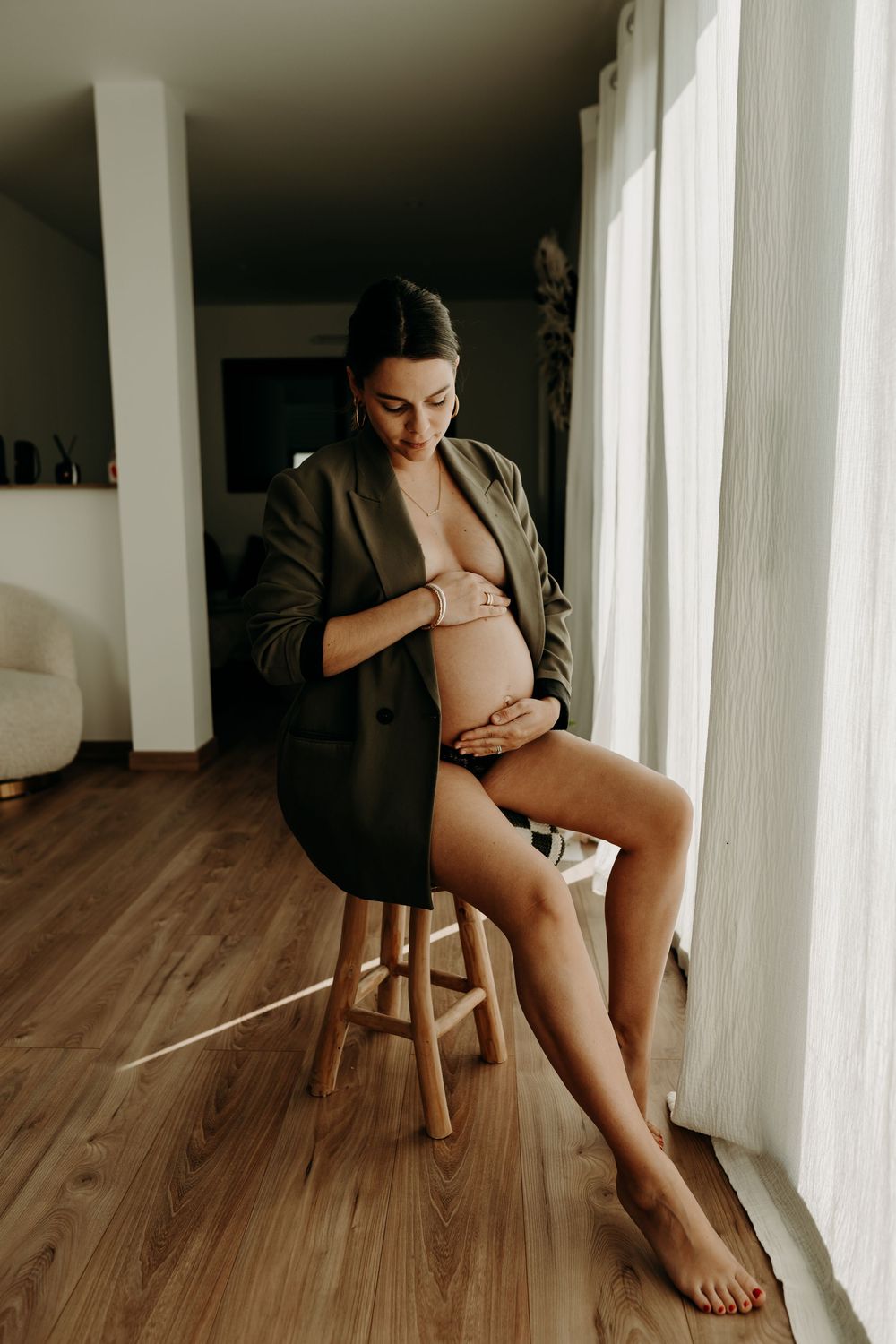 Artistic maternity photo showing an intimate moment by a window, with soft natural light on hardwood floors.