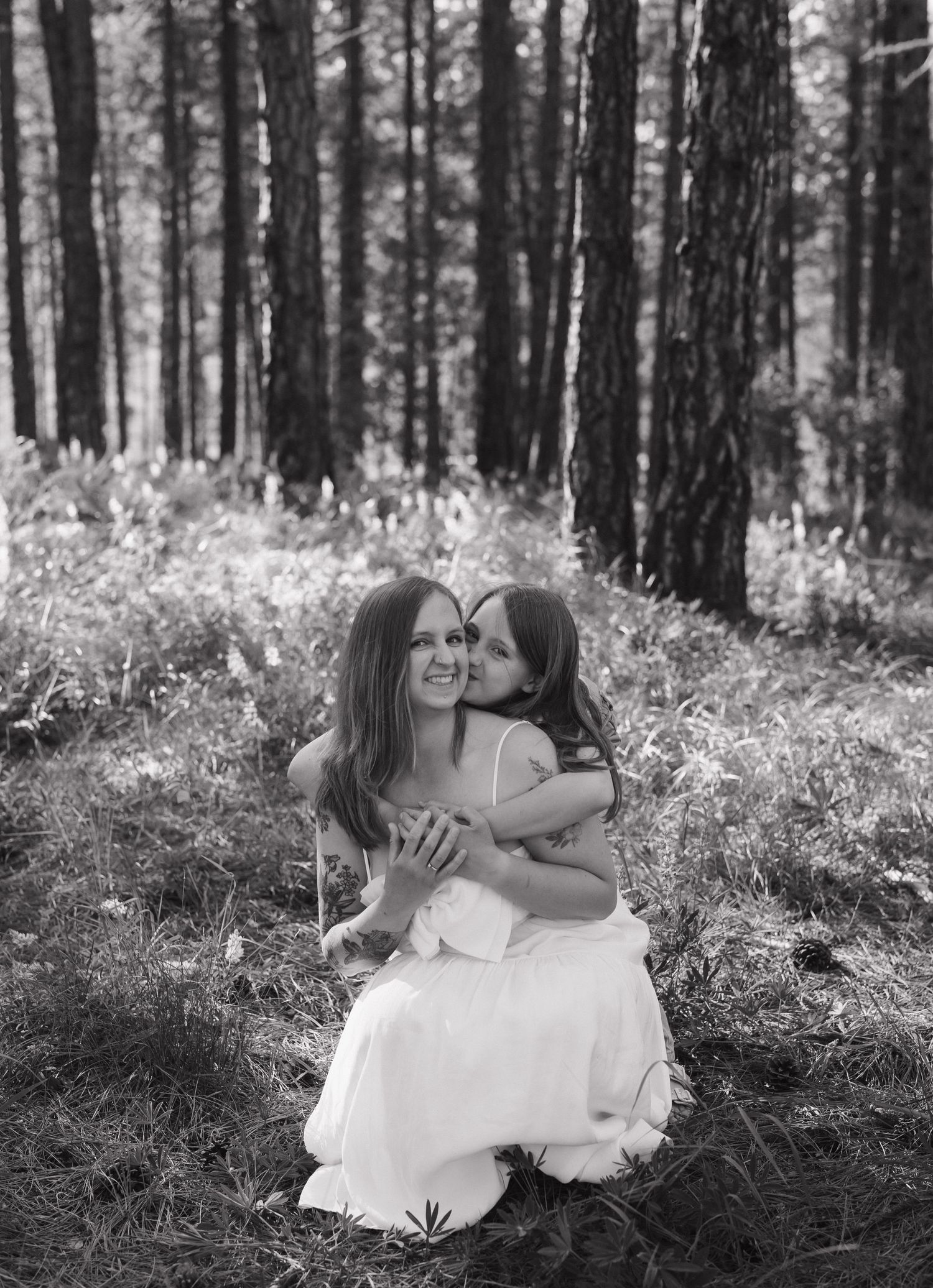 Black and white photo of a mother and young daughter embracing in a sunlit forest, both wearing white dresses.