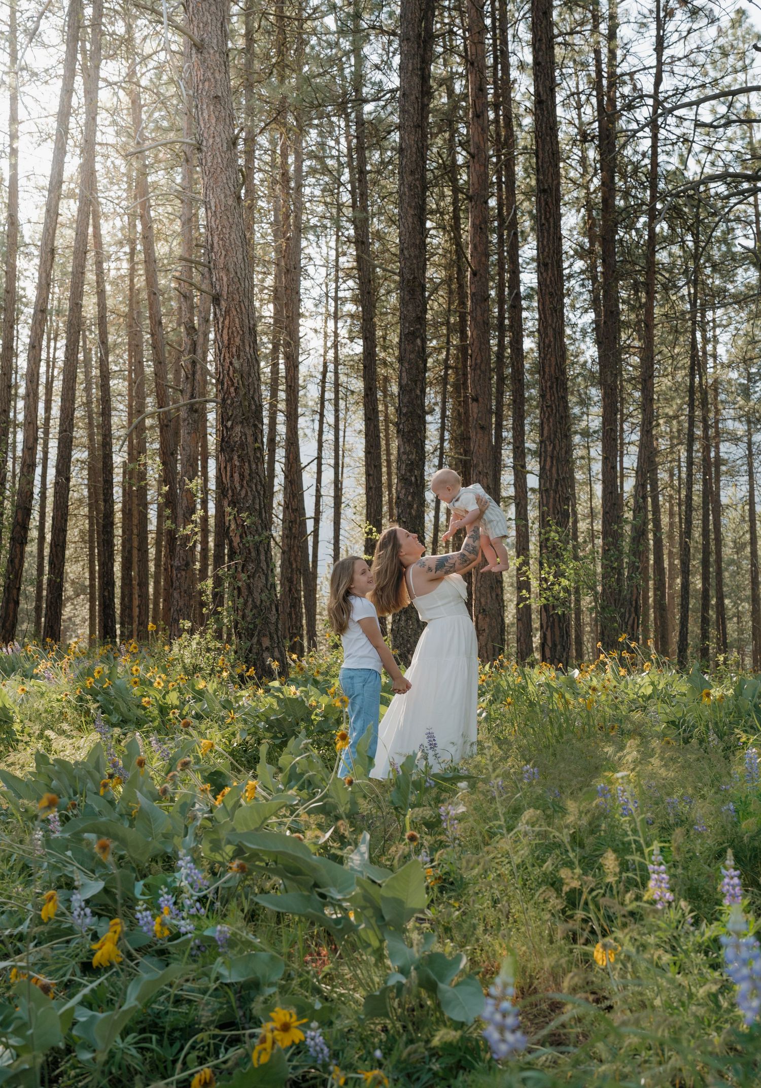 Family with young children playing in a sunlit forest meadow filled with wildflowers among tall pine trees.