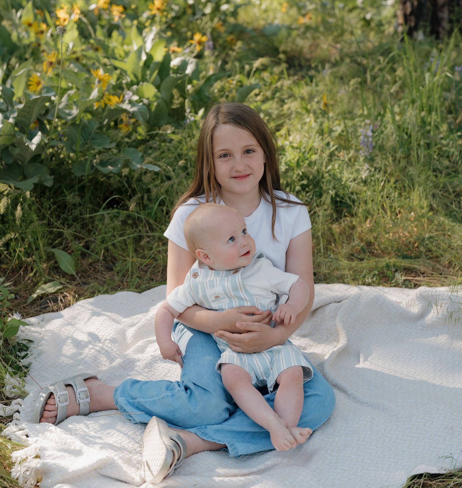 Young girl sitting on a blanket outdoors holding a baby sibling, surrounded by lush green grass and yellow wildflowers.