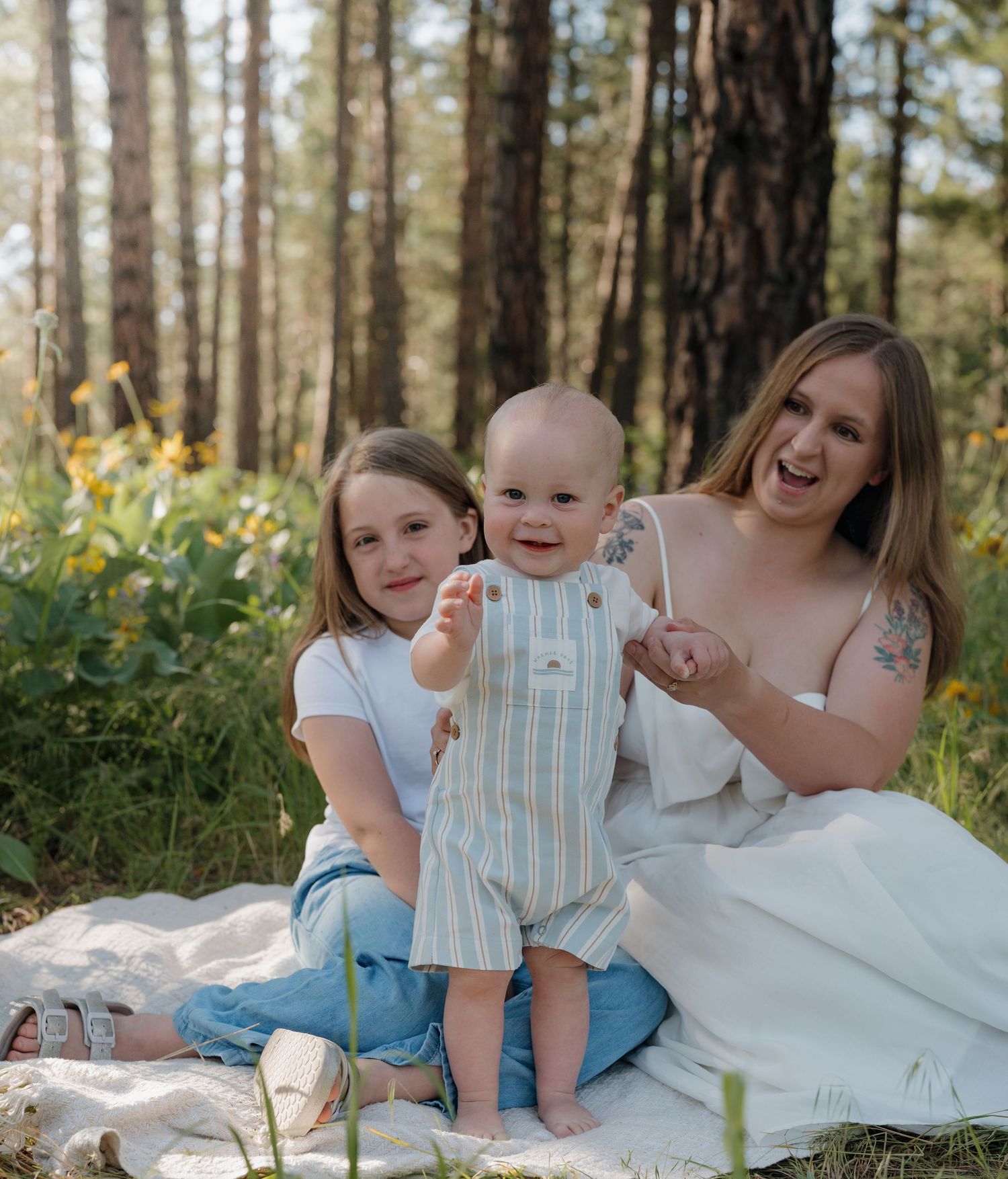 A young mother sits on a blanket in a sunny forest with her two children, a girl and a smiling baby in a striped outfit.