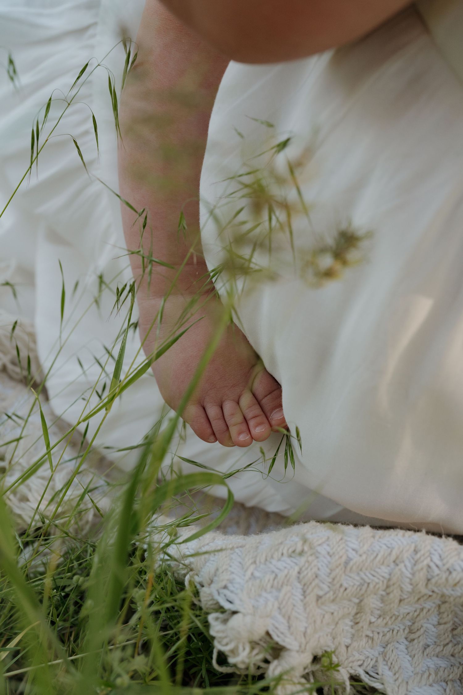 Bare feet of a person in a white dress sitting in tall green grass outdoors.