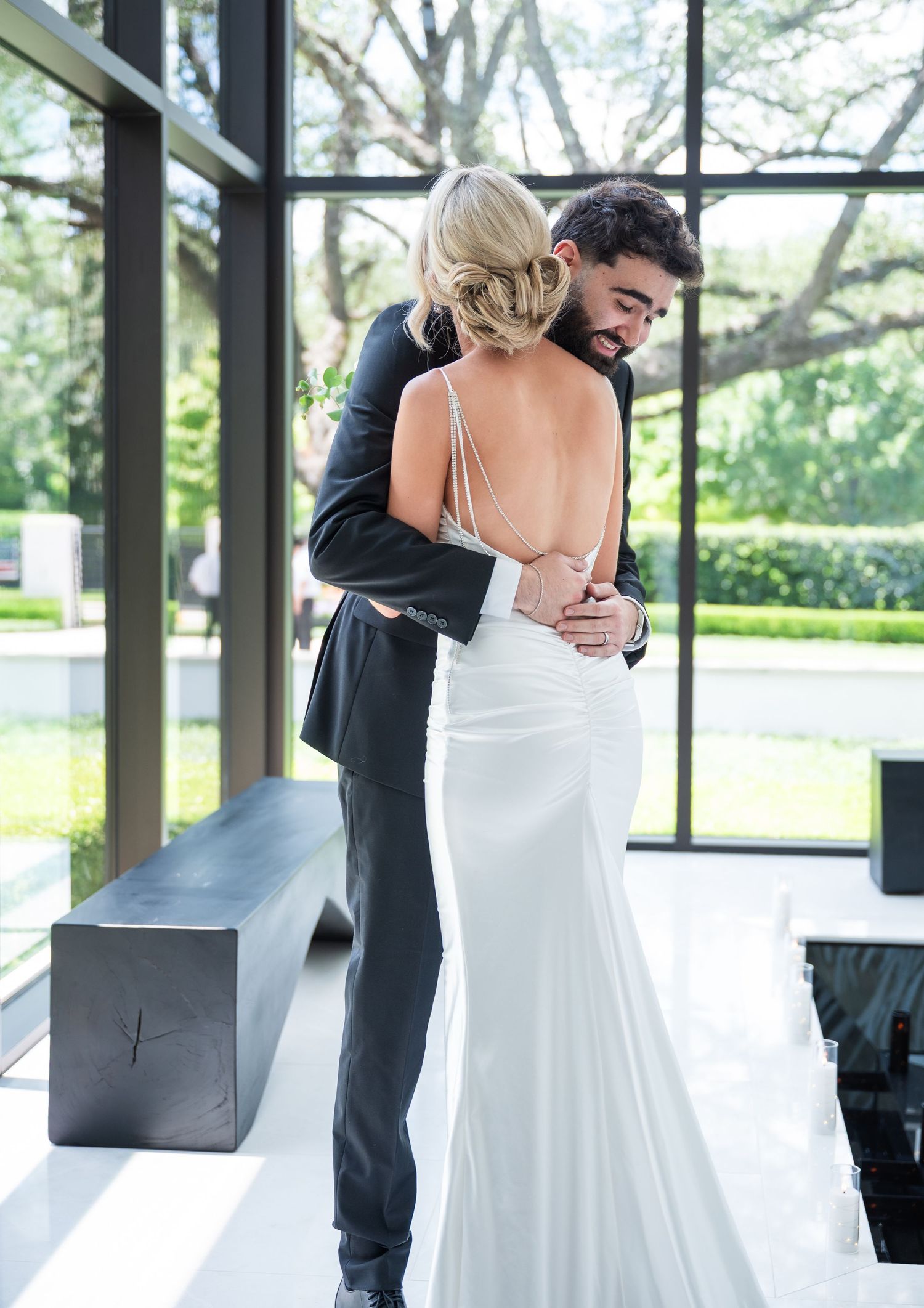 A bride in a backless white gown embraces her groom in a black suit near large windows with garden views.