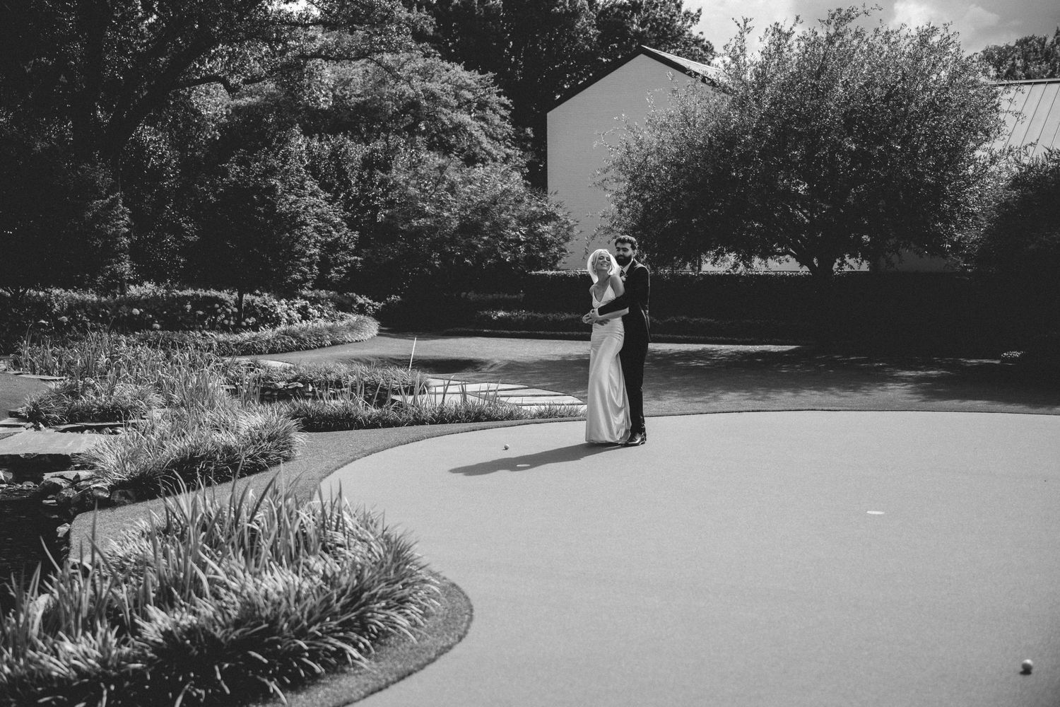 Black and white photo of a couple embracing in a garden path near a pond on their wedding day.