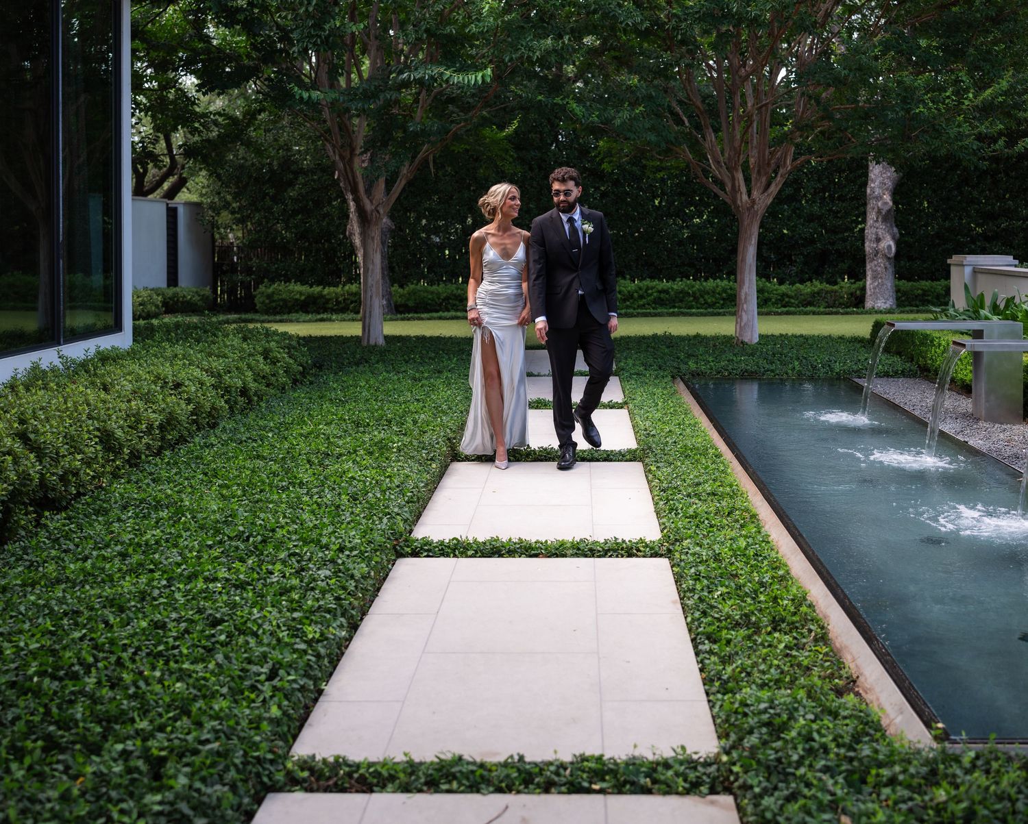 A bride and groom walk hand in hand along a stone path surrounded by lush green hedges and a reflecting pool.