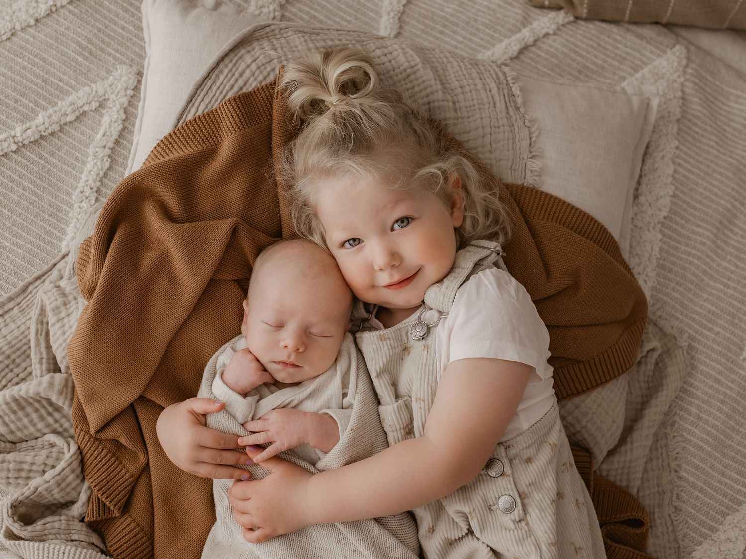 Siblings bonding moment with older sister holding newborn on cream textured bedding.