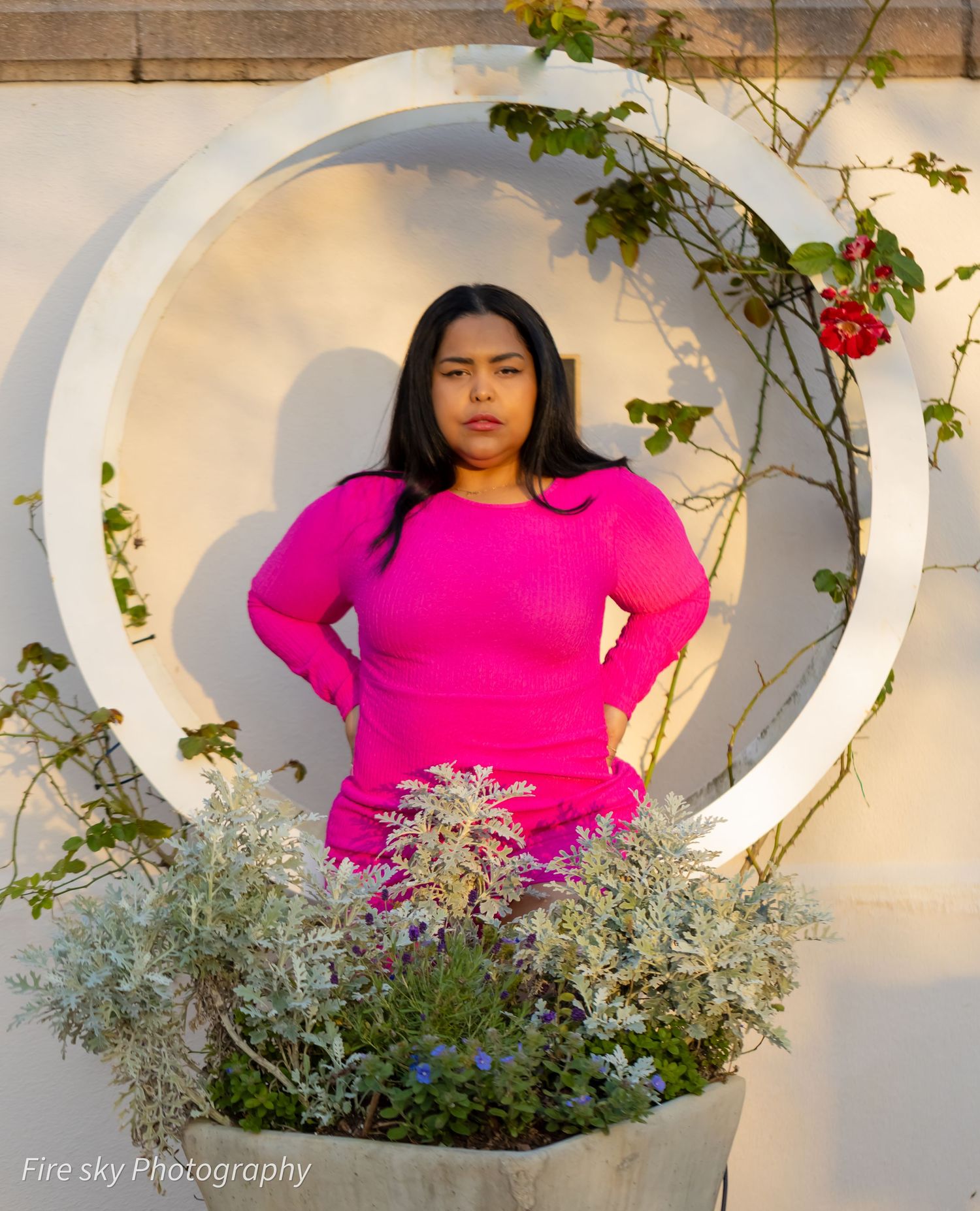 A person in a bright pink sweater poses in front of a circular architectural feature with flowering vines.