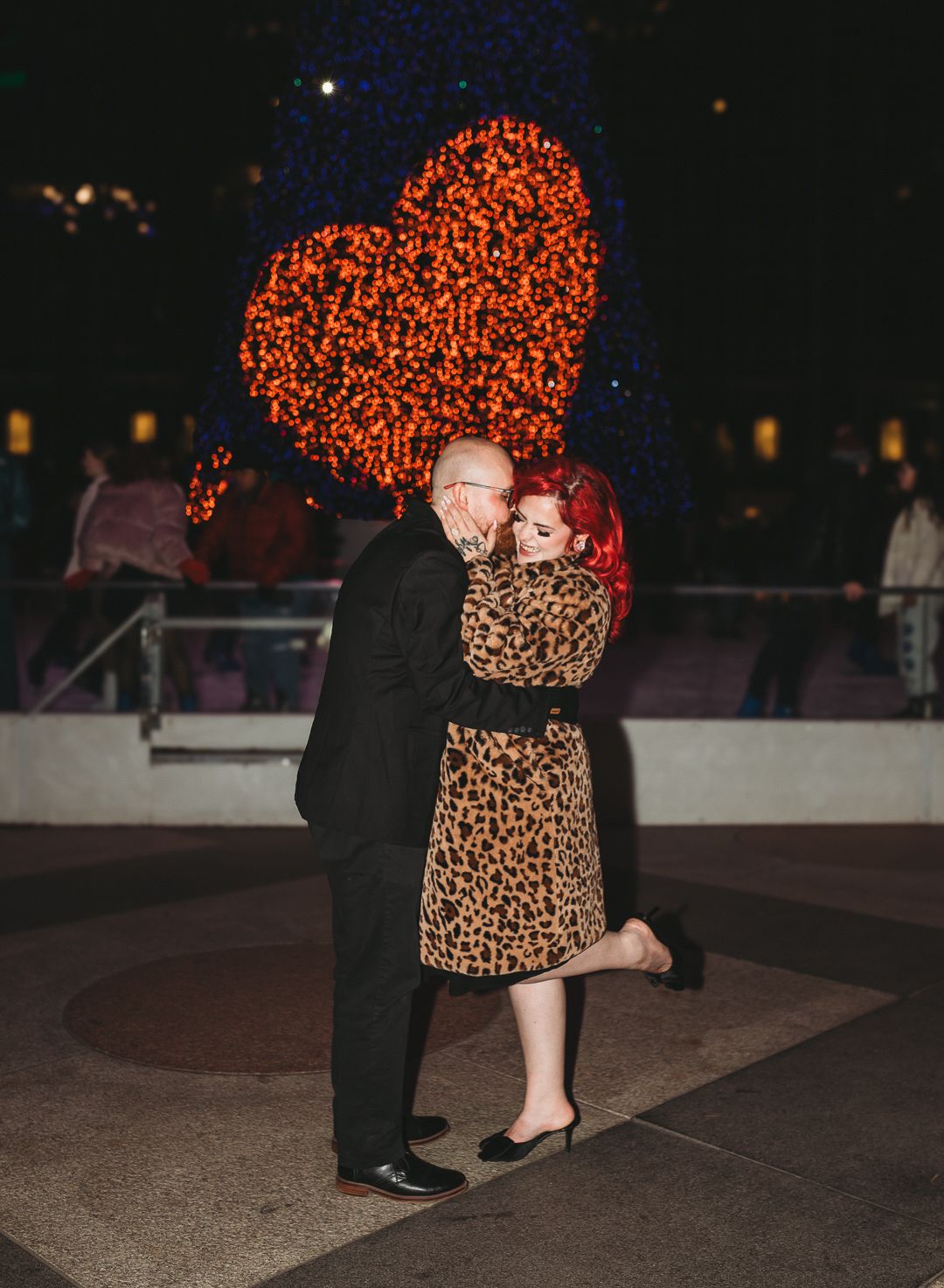 A couple embraces under a glowing red heart display at night. Pittsburgh Engagement Photographer