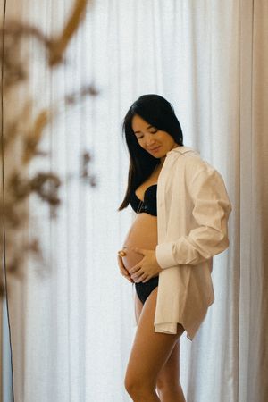 Woman in white shirt poses against curtained window in artistic boudoir photoshoot.