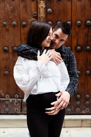 A couple in romantic embrace wearing black and white outfits pose against a rustic wooden door background.