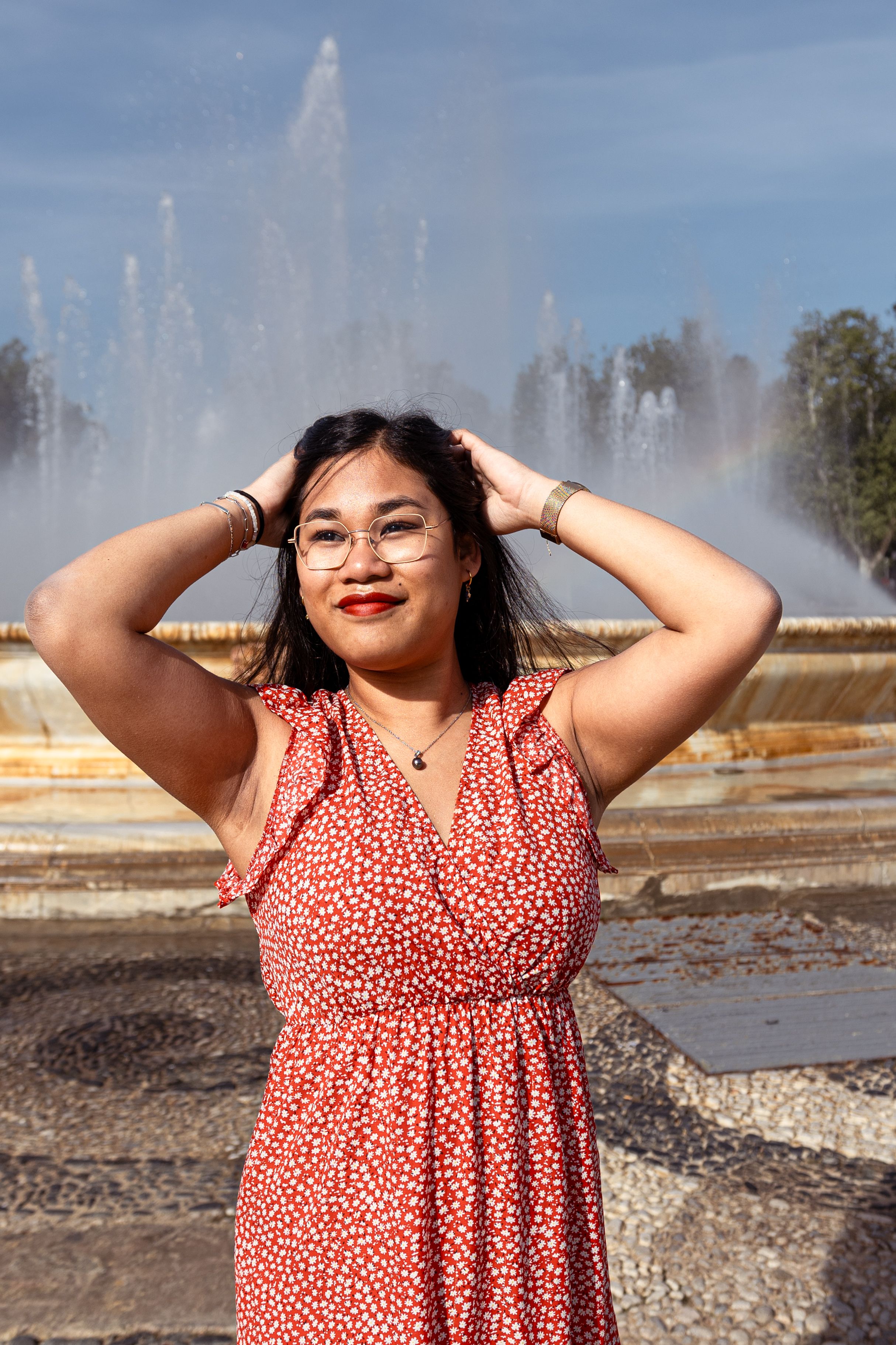 Woman in a coral polka dot dress poses joyfully in front of a fountain on a sunny day.  shooting de femme séville