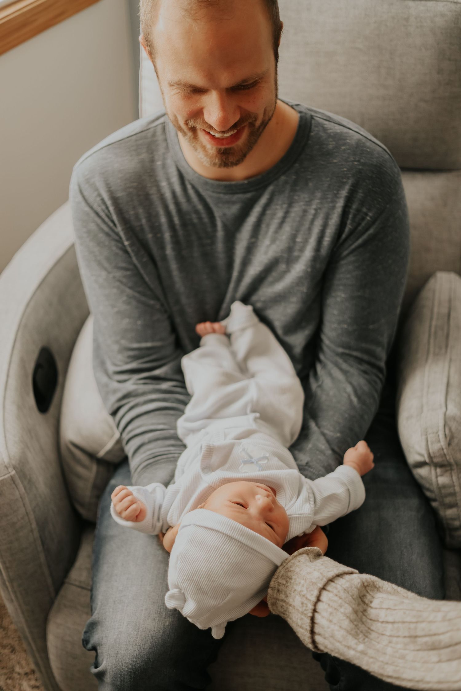 Parent sitting in grey armchair sharing joyful moment with infant in white onesie.