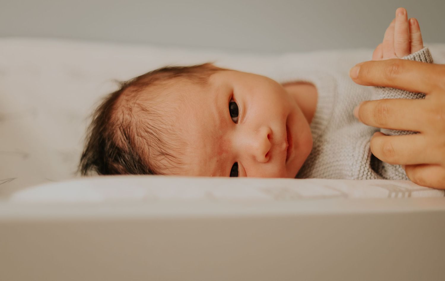 A newborn baby lies on white bedding while looking up at the camera with curious eyes.